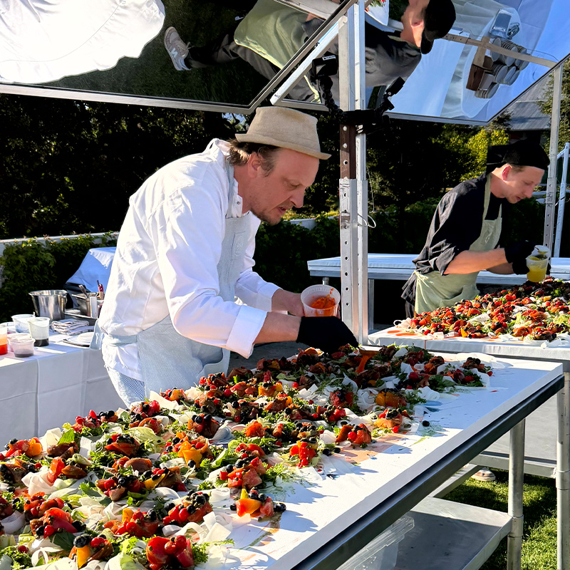 Two chefs prepare a colorful spread of vegetable dish on a long outdoor table.