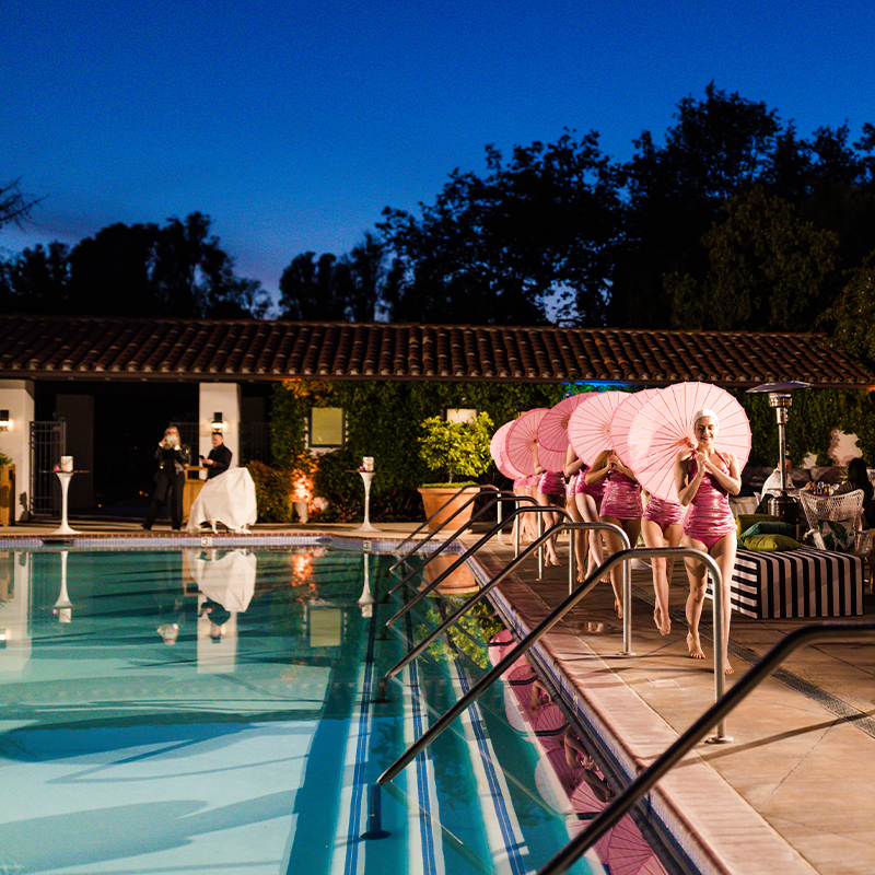Evening outdoor poolside event with performers in pink swimsuits carrying parasols along a lit deck, their colorful reflections shimmering in calm blue water.