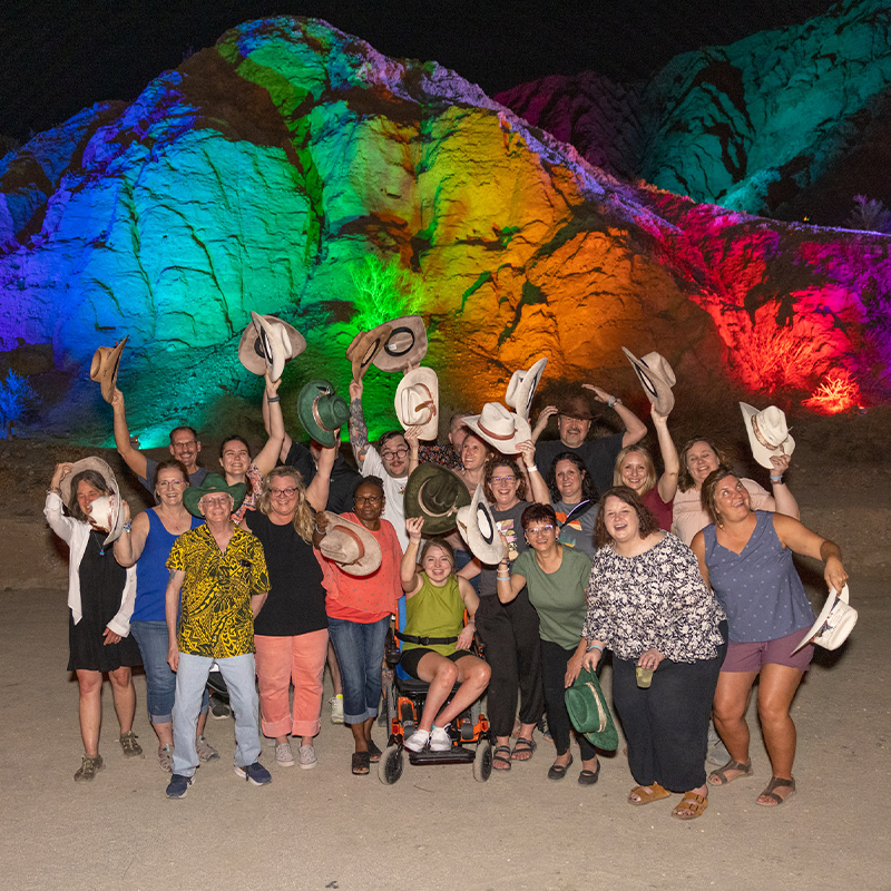 group of employees smile and pose with cowboy hats in front of rainbow projection over a Palm Springs landscape