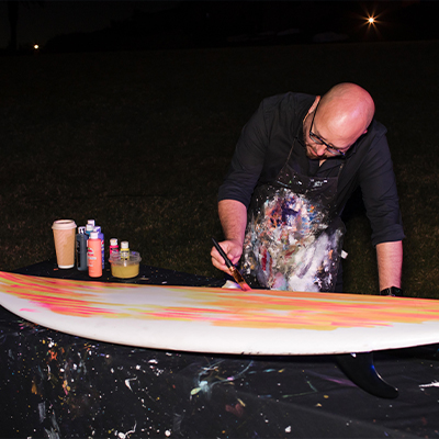 Artist paints a surfboard in pinks and oranges at Los Angeles event