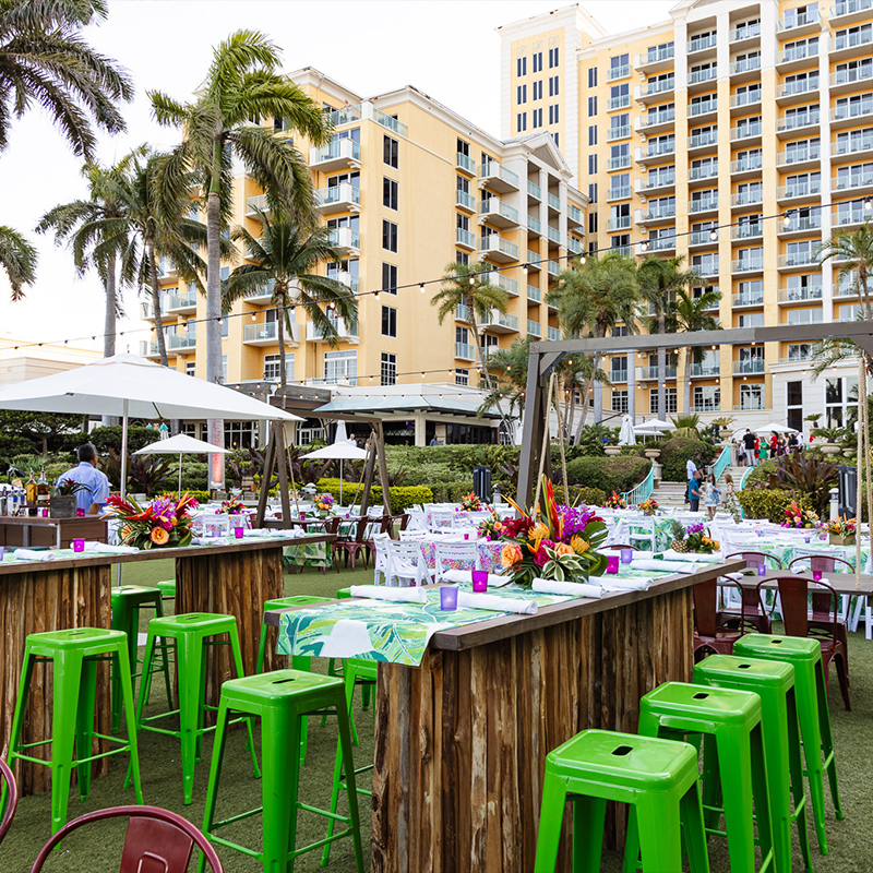 Outdoor dining area with vivid green stools, flower arrangements, and white-clothed tables beneath umbrellas, framed by palm trees