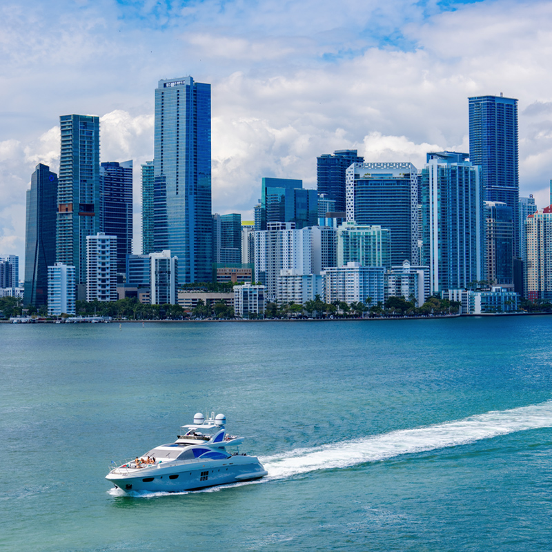 white-and-blue yacht speeds across turquoise waterfront, leaving a foamy wake before a dense skyline of glass towers under a cloudy blue sky of Miami