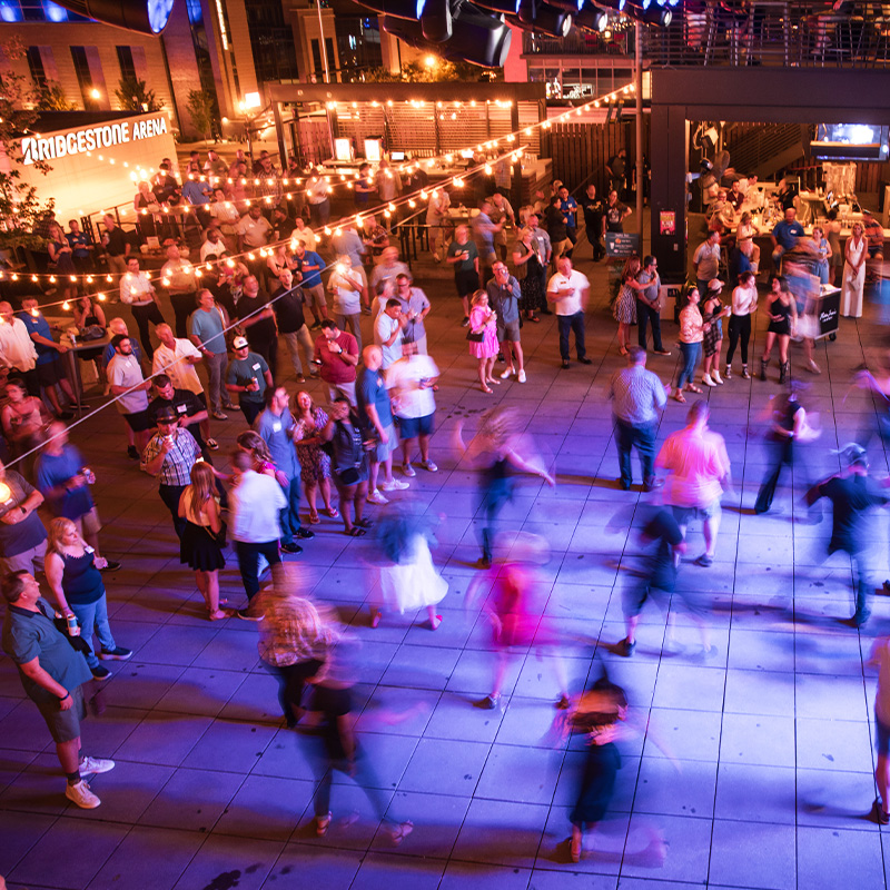 lively outdoor dance gathering near Bridgestone Arena, with glowing string lights, watching crowds, and dancers blurred in colorful blue and pink light