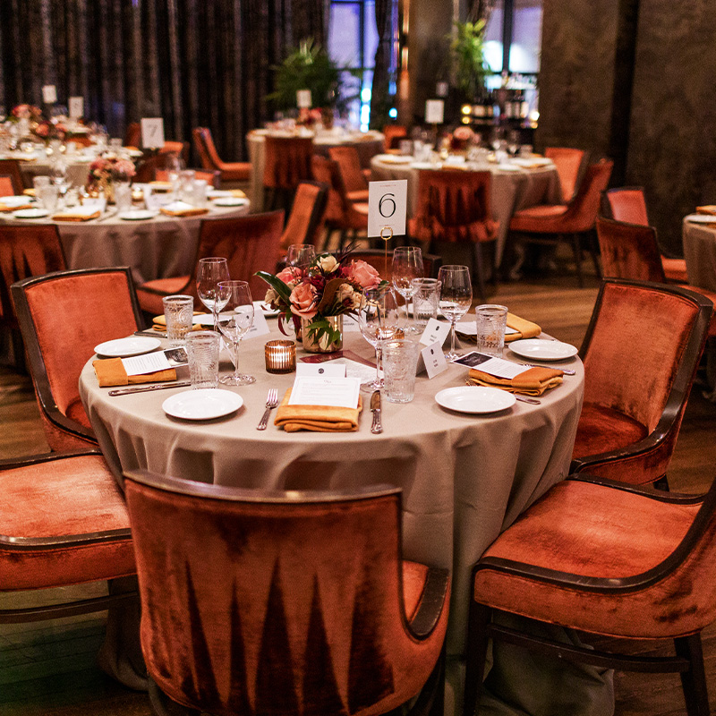 An elegant banquet hall features round tables with taupe linens, floral centerpieces, glassware, and orange place settings, surrounded by plush rust-red chairs.