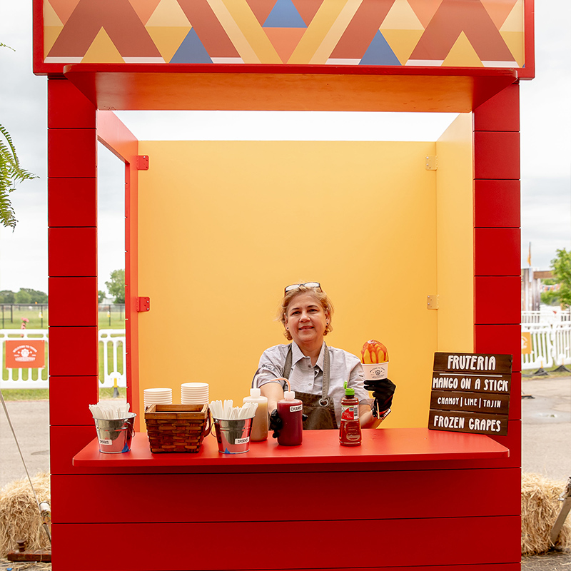 A smiling vendor stands in a bright red-and-yellow Tex-Mex food kiosk, holding a frozen treat beside cups and toppings