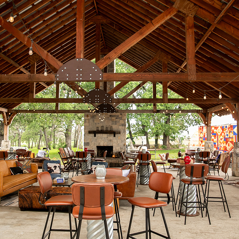 An airy open-sided pavilion with a high timber-beamed roof, stone fireplace, and scattered tables and rust-colored chairs