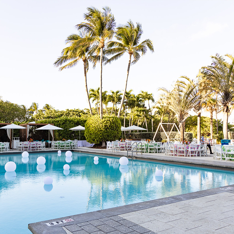 A tranquil resort pool with floating white balls, lounge chairs, dining tables and umbrellas, tall palm trees, and a stone deck in bright sunshine on a beackfront