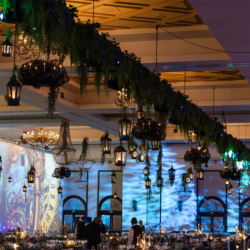 A grand banquet hall glows with chandeliers and hanging lanterns beneath a canopy of greenery