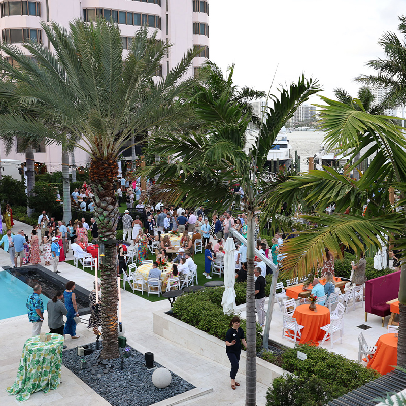 A lively outdoor waterfront reception beneath palm trees, with guests mingling around white and orange tables