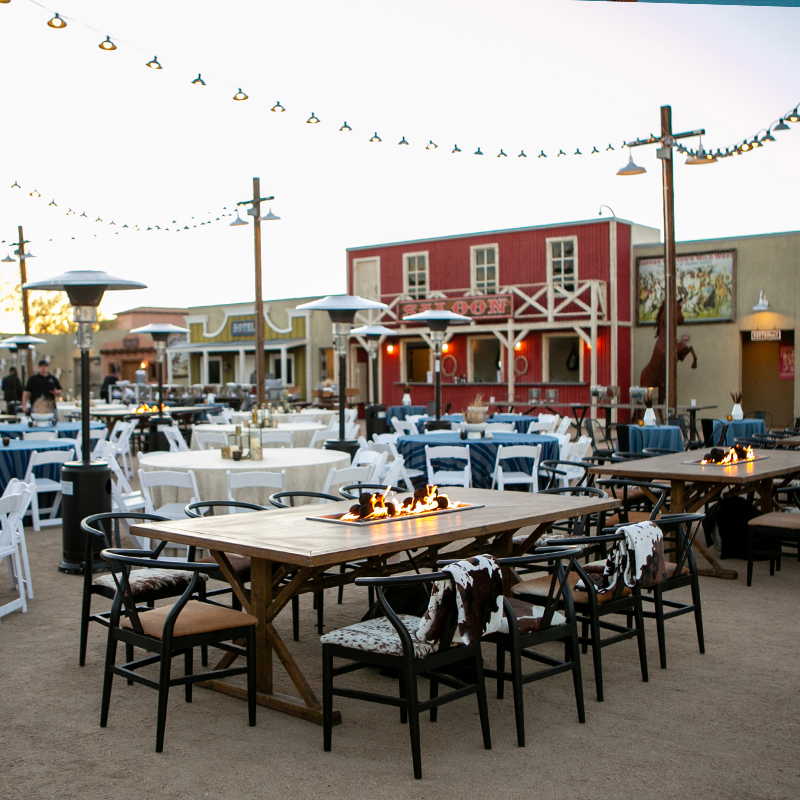 outdoor set up of rustic tables, white chairs, tabletop flames, and overhead string lights, facing a red barn-style storefront