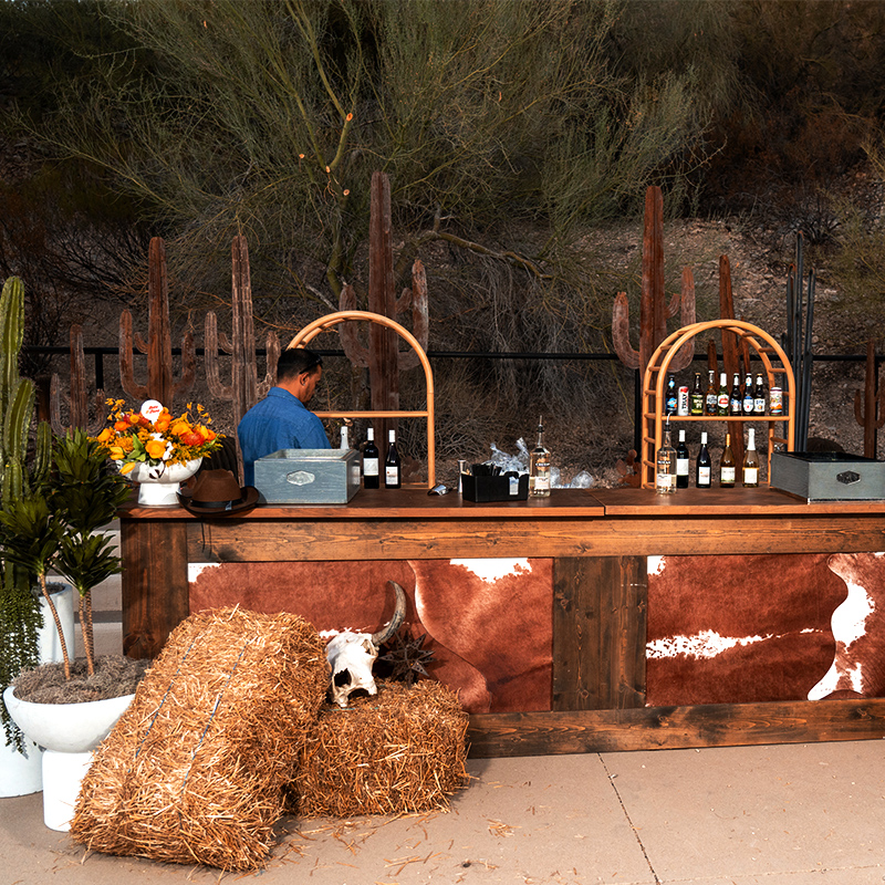 An outdoor rustic bar with a bartender, bottles, hay bales, and a cowhide-fronted counter, set among tall cacti and desert brush