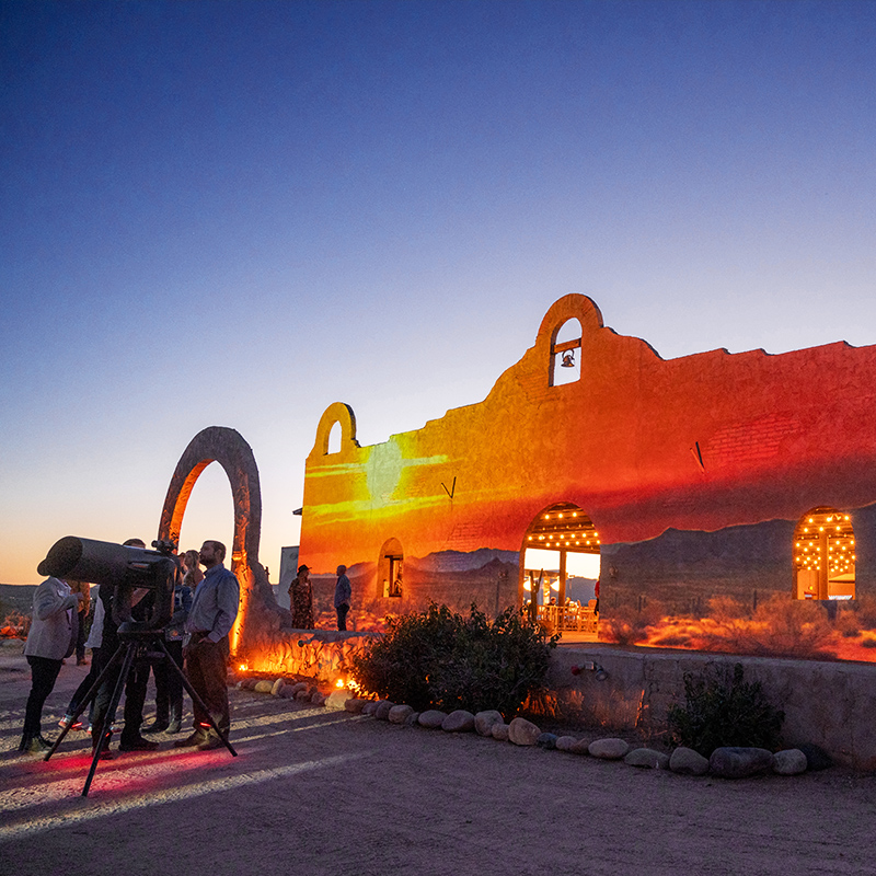 Visitors gather around a telescope beside a glowing desert adobe building at sunset