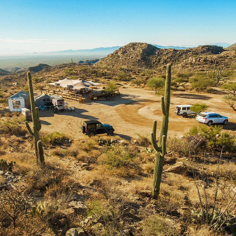 a desert campsite that sits among rocky hills and tall saguaro cacti, with tents, a rustic shelter, and parked vehicles beneath a clear blue sky